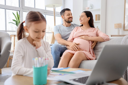 Happy pregnant couple sitting on sofa while their little daughter drawing at homeの写真素材