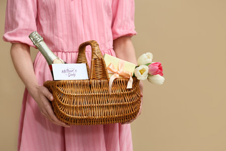 Young woman holding basket with tulips, champagne bottle and gift box on beige backgroundの写真素材