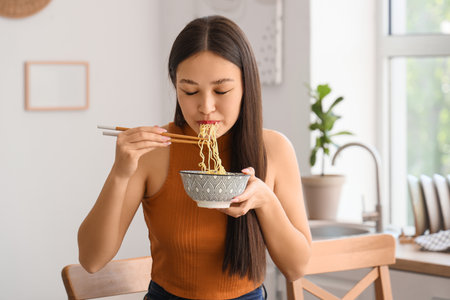 Young Asian woman eating tasty ramen in kitchenの写真素材