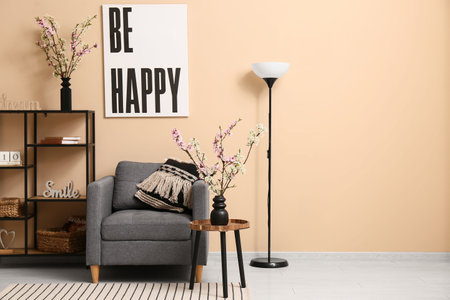 Armchair with shelving unit, coffee table and vases of blooming branches near beige wall in living roomの写真素材