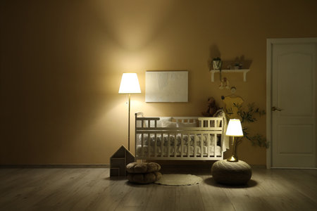 Interior of children's bedroom with baby crib, shelf and glowing lamps near beige wall at nightの写真素材