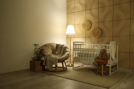Interior of children's bedroom with baby crib, armchair and glowing lamp near tile wall at nightの写真素材