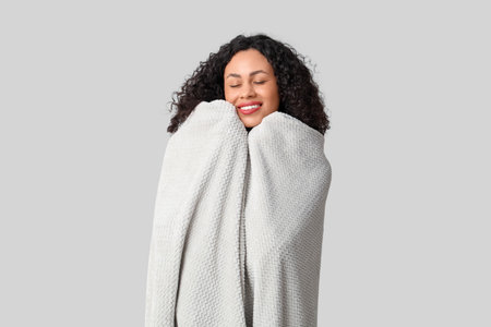 Beautiful young African-American woman with cozy blanket on gray backgroundの写真素材