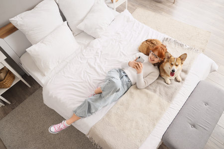 Teenage redhead girl with cute Corgi dog using mobile phone in bedroom, top viewの写真素材