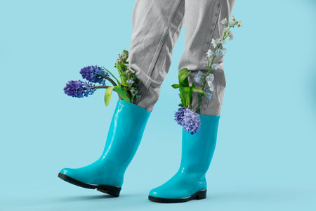 Legs of young woman in rubber rain boots with beautiful flowers on blue background