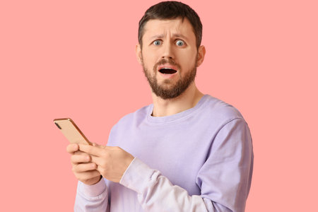 Shocked young man using mobile phone on pink backgroundの写真素材