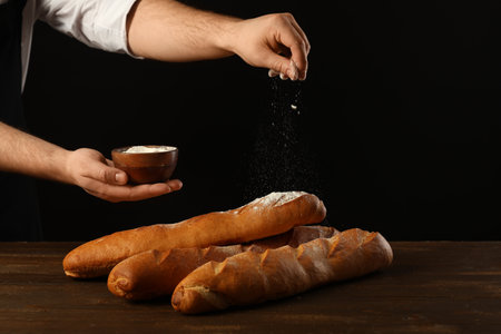 Man sprinkling flour on fresh baguettes against black backgroundの写真素材