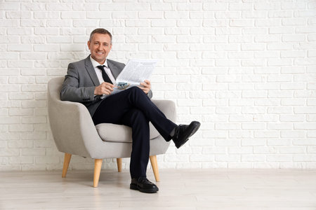 Portrait of mature businessman with modern laptop sitting on armchair near white brick wallの写真素材
