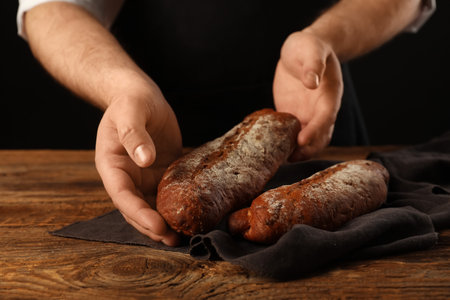 Man with fresh rye baguettes on wooden table against black background, closeup