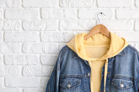 Hanger with stylish yellow hoodie and jeans jacket on white brick backgroundの写真素材