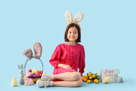 Cute Asian girl in bunny ears with Easter decor sitting on blue backgroundの写真素材