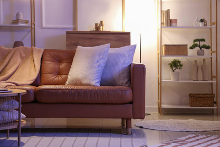 Interior of living room with brown sofa, shelf unit and glowing lamp in eveningの写真素材