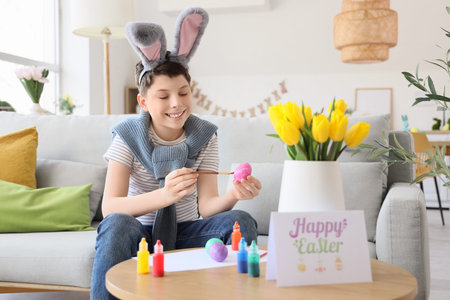 Teenage boy in bunny ears painting Easter egg on sofa at home