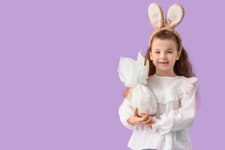 Cute little girl in bunny ears with Easter gift egg on lilac background