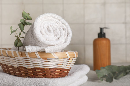 Wicker basket with clean towels, cosmetic product and eucalyptus branches on table against light tile wallの写真素材