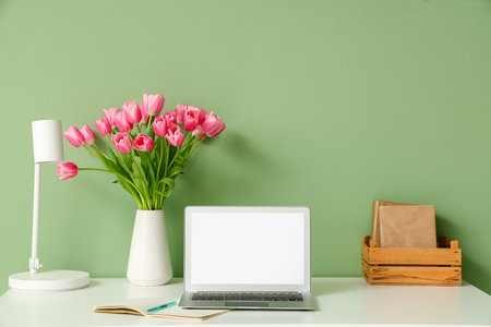 Stylish workspace with modern laptop, books and bouquet of beautiful tulip flowers near green wallの写真素材