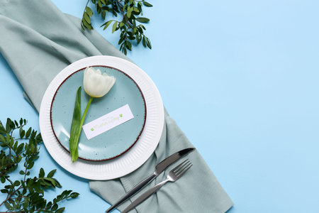 Beautiful table setting with greeting card and tulip flower on blue background. Mother's Day...