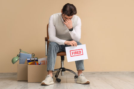 Upset young man holding paper with the word FIRED in chair near beige wallの写真素材