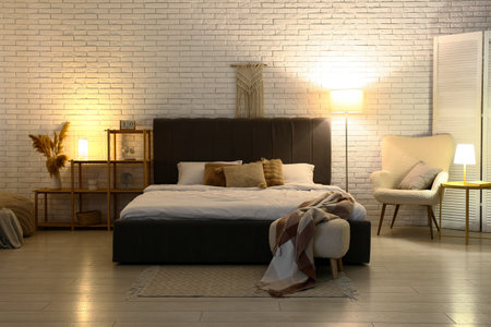 Stylish interior of bedroom with soft gray bed, comfortable armchair and glowing lamps at nightの写真素材