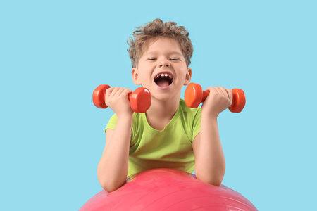 Sporty little boy exercising with heavy dumbbells on fitball against blue backgroundの写真素材