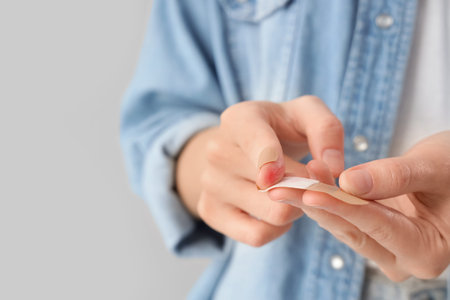 Young woman applying medical plaster onto injured finger against gray background, closeup