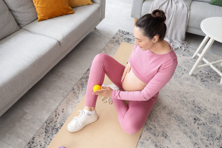Sporty pregnant woman massaging her leg with ball on mat at home