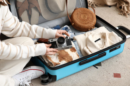Young woman unpacking her suitcase with clothes and photo camera at home, closeup