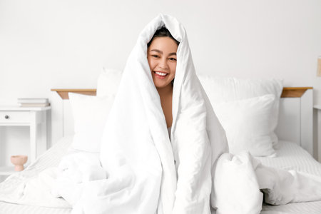 Smiling young woman with blanket sitting in bedroomの写真素材