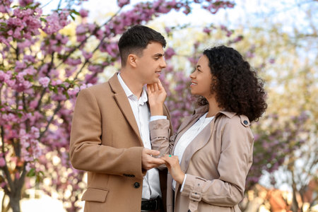 Beautiful young happy couple looking at each other near blooming trees in park on spring dayの写真素材