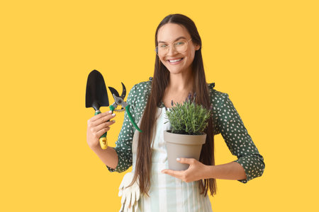 Beautiful young woman holding pot with lavender flowers and gardening tools on yellow backgroundの写真素材