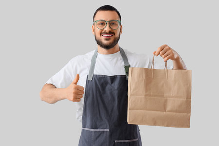 Male worker with paper bag showing thumb-up on light background. Food delivery serviceの写真素材