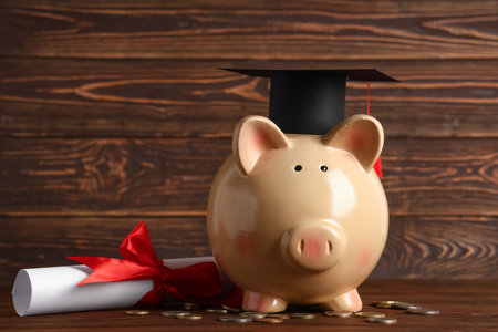 Piggy bank with graduation hat, diploma and money on table against wooden background. Tuition fees conceptの写真素材