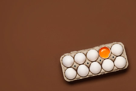 Broken chicken egg among whole ones in cardboard holder on color backgroundの写真素材