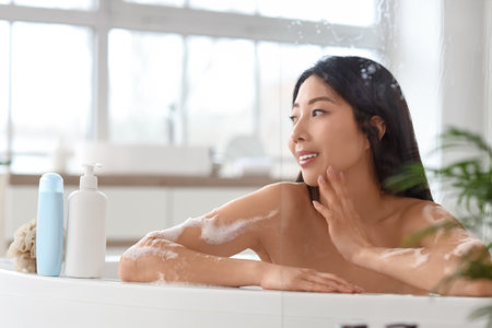 Pretty young Asian woman with bottles of shower gel and loofah taking bath in bathroomの写真素材