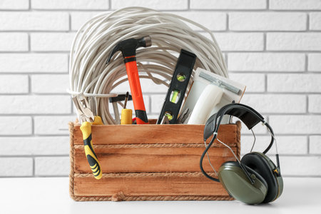 Box with protective headset and construction tools on table against white brick background