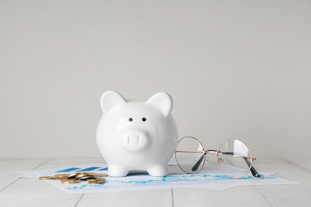 Piggy bank with coins, eyeglasses and diagrams on white tile tableの写真素材