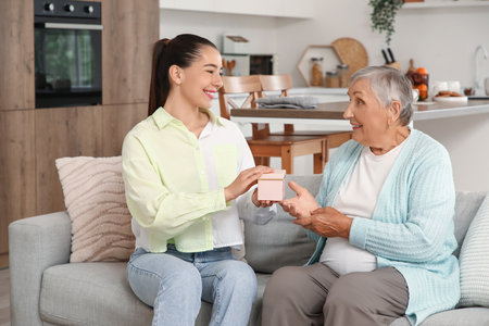 Young woman greeting her grandmother with gift on sofa at homeの写真素材