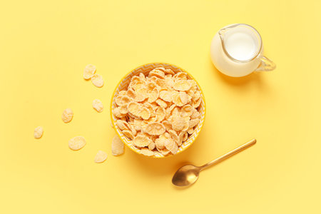Composition with bowl of tasty cornflakes and jug of milk on yellow background