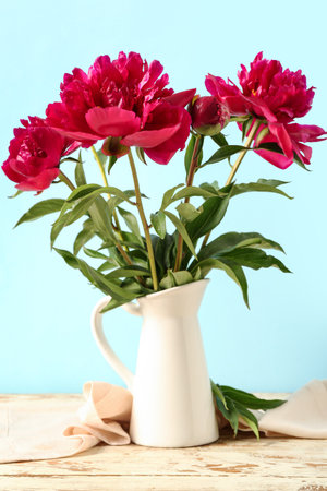 Jug with beautiful peony flowers on white wooden table against blue backgroundの写真素材
