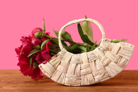 Wicker bag with beautiful peony flowers on wooden table against pink backgroundの写真素材