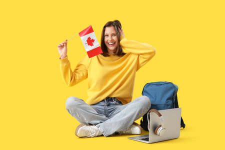 Female student with flag of Canada and backpack sitting on yellow backgroundの写真素材