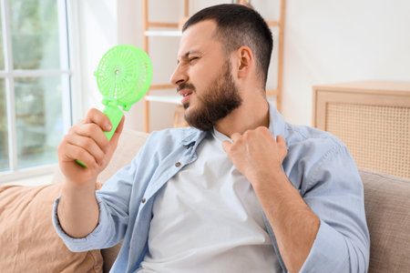 Young man with mini fan suffering from heat on sofa at homeの写真素材