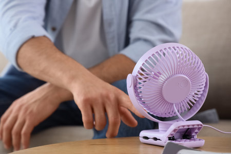 Young man turning mini fan on table at home, closeup