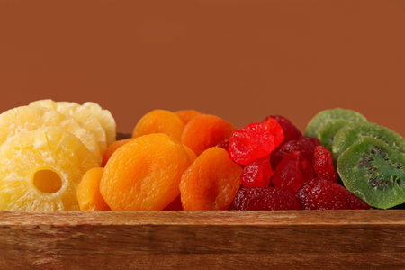 Wooden tray with different dried fruits on brown background, closeup