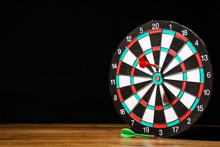 Board with darts on wooden table against dark background