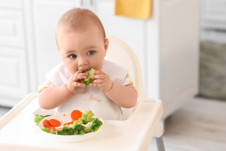 Cute little baby eating broccoli in high chair at homeの写真素材