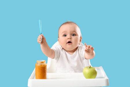 Cute little baby with puree and apple in high chair on blue backgroundの写真素材