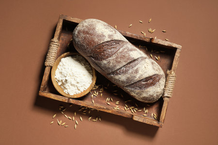 Tray with loaf of fresh rye bread, wheat and flour on brown backgroundの写真素材