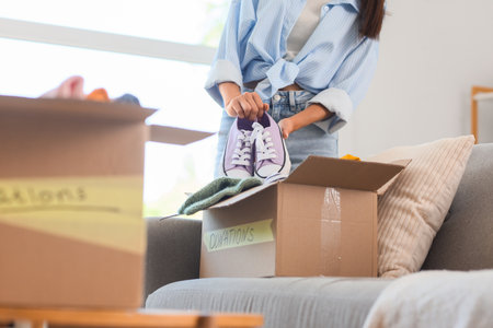 Young woman packing sneakers into box for donation on sofa at homeの写真素材