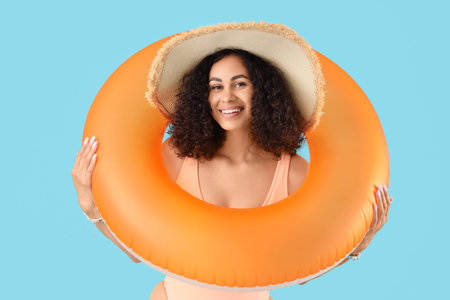 Young African-American woman in swimsuit with orange inflatable ring on blue background